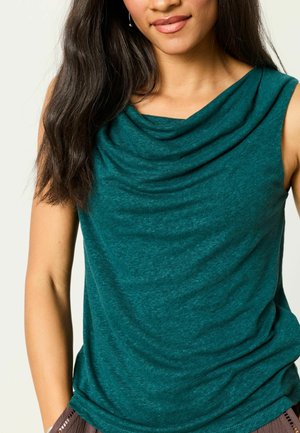 Frau mit langem dunklem Haar, die ein ärmelloses türkisfarbenes Oberteil mit drapiertem Ausschnitt und Ohrringen trägt, eine Hand teilweise in der Tasche.