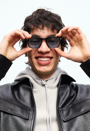 Mladý člověk se usmívá a nastavuje si černé sluneční brýle, nosí šedou mikinu pod černou koženou bundu na bílém pozadí.