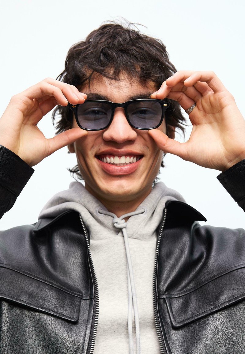 Jeune personne souriante ajustant des lunettes de soleil noires, portant un sweat à capuche gris sous une veste en cuir noire sur un fond blanc.