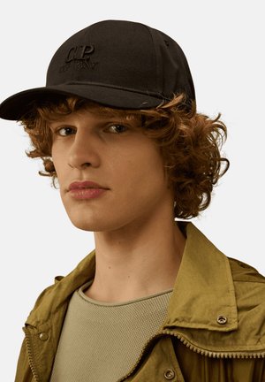 Giovane con capelli ricci che indossa un berretto da baseball nero C.P. Company, giacca verde oliva e camicia chiara dalla texture leggera, rivolto verso la fotocamera su sfondo uniforme.