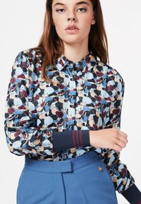 Bluse mit Blumenmuster, die blaue, burgunderfarbene und cremefarbene Blumen auf schwarzem Hintergrund zeigt. Sie verfügt über einen geknöpften Kragen und gerippte marineblaue Bündchen mit Streifen.