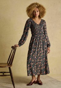 Langes Blumenkleid mit dunklem Hintergrund, das lange Ärmel, ein gerafftes Oberteil und einen fließenden Rock hat. Getragen mit burgunderfarbenen High Heels.