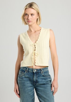 Top corto sin mangas de color crema con un escote ondulado, un patrón de tejido texturizado y un detalle de lazo en la parte frontal. Combinado con unos jeans de mezclilla azul.