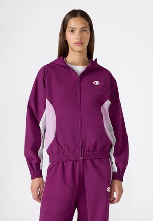 Joven mujer que lleva una chaqueta con capucha y cremallera en púrpura y rosa y pantalones púrpuras a juego con un pequeño logotipo en el pecho y el muslo.