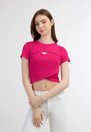Tætsiddende lyserød ribbet crop top med korte ærmer og et krydset design foran, som afslører et lille maveområde. Båret med hvide bukser.