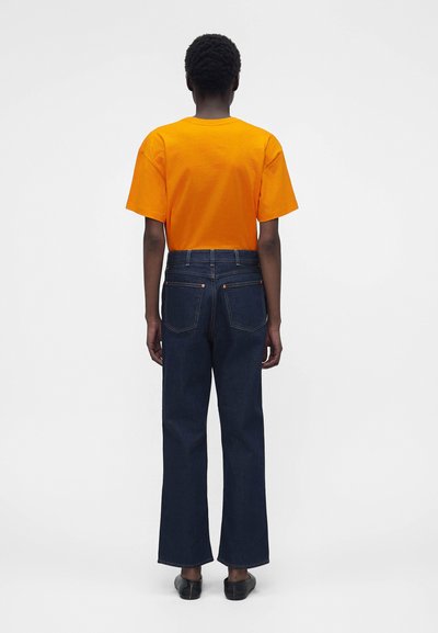 Personne aux cheveux courts et bouclés, debout de dos, portant un t-shirt orange à manches courtes, un jean bleu foncé et des chaussures noires sur un fond blanc.