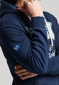 Felpa con cappuccio blu navy realizzata in morbido tessuto, con logo frontale e cordini. Include un'etichetta blu sulla manica sinistra e polsini a coste.