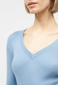 Femme portant un pull léger bleu clair à côtes avec col en V et manches longues, visible des épaules jusqu'au milieu de la poitrine sur un fond uni clair.
