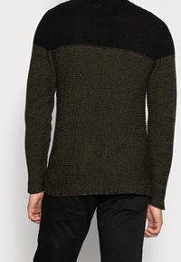 Mörkgrön och svart stickad tröja med texturerat tyg, långa ärmar och rundad nederkant. Bakifrån med kontrasterande axeldesign.