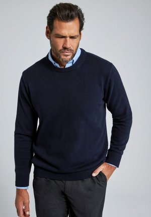 Maglione lavorato a maglia blu navy con colletto rotondo e polsini a coste, indossato sopra una camicia colletto blu chiaro, abbinato a pantaloni scuri.