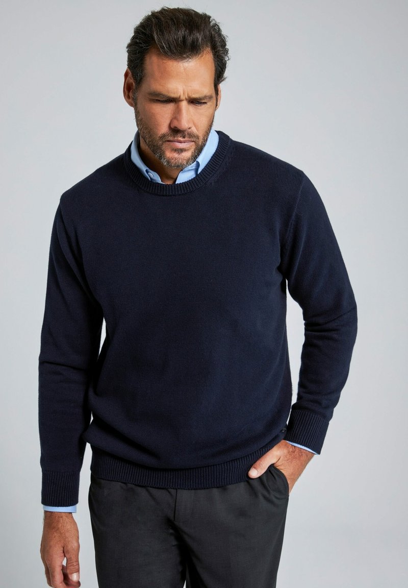 Maglione lavorato a maglia blu navy con colletto rotondo e polsini a coste, indossato sopra una camicia colletto blu chiaro, abbinato a pantaloni scuri.
