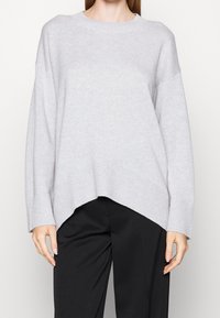 Pull gris clair oversize avec texture côtelée, col rond et ourlet asymétrique. Associé à un pantalon noir pour créer du contraste.