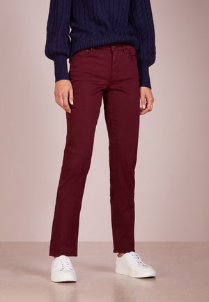 Osoba nosząca ciemnofioletowe spodnie typu slim fit, granatowy sweter w warkocze oraz białe trampki, stojąca na jasnej podłodze na jednolitym tle.
