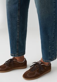 Brown suede low-top sneakers with dark brown accents, rubber sole, and rounded laces. Worn with blue straight-leg jeans.