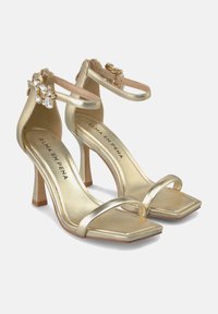 Goldene, metallic glänzende High-Heelsandalen mit eckiger Zehenpartie, dünnen Knöchelriemen und dekorativen Kristallakzenten. Die Sohle ist hellbraun.