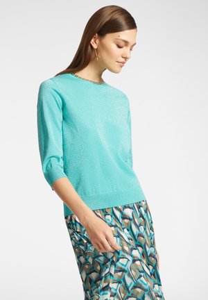 Maglione in maglia metallizzata turchese con maniche a tre quarti, orlo a coste e colletto rotondo, abbinato a una gonna con fantasia.