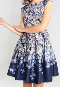 Blumenmuster-Kleid mit tailliertem Oberteil, kurzen Ärmeln und schwingendem Rock. Der Stoff ist navyblau mit bunten Blumenmustern durchzogen.