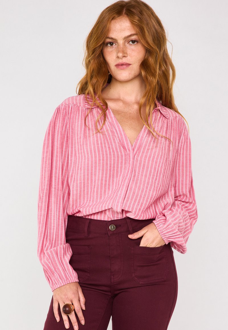 Blusa a righe rosa con maniche lunghe e ampie e scollatura a V, abbinata a pantaloni attillati bordeaux scuro con tasche frontali.