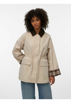 Göndör hajú nő bézs dzsekiben, mely sötét gallérral, kockás mandzsettákkal és elülső zsebfedőkkel rendelkezik, kék farmerrel párosítva.