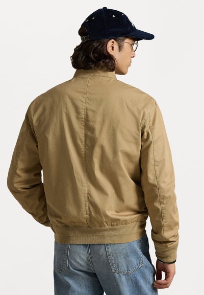 Braune Bomberjacke aus Baumwolle, mit einem gerippten Bund, glatter Textur und einfachem Design. Getragen mit hellblauen Jeans und einer Kappe.