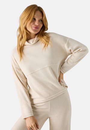 Sudadera beige ligera con cuello de capa, mangas largas y un corte ligeramente holgado; combinada con pantalones ligeros a juego. Textura suave.