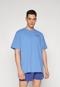 Tricou din bumbac bleu deschis cu mâneci scurte, guler rotund și un logo negru pe partea stângă a pieptului. Modelul poartă pantaloni scurți albaștri.
