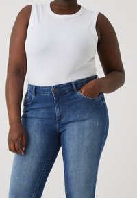 Vit ribbad tanktop kombinerad med blå denimjeans. Jeansens design är klassisk med fem fickor och knappstängning framtill.