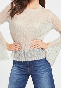 Knit sweater in light beige with vertical stripes, feature bell sleeves, and a textured finish. Paired with blue denim jeans.