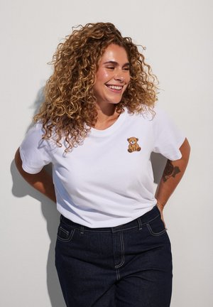 Femme aux cheveux bouclés souriante, portant un t-shirt blanc avec un petit écusson nounours et un jean foncé, debout devant un fond uni.