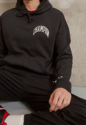 Felpa nera con logo "Champion" in caratteri bianchi. Caratterizzata da cappuccio con coulisse, tasca a marsupio e maniche con polsini. Abbinata a pantaloni neri.