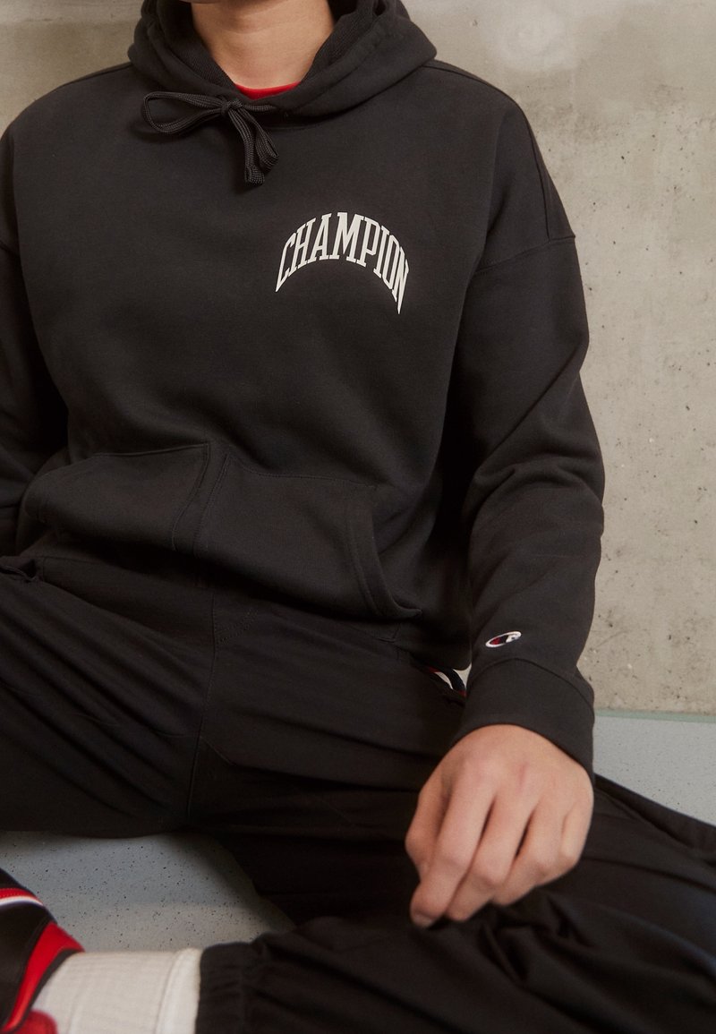 Black hoodie with "Champion" logo in white font. Features a drawstring hood, kangaroo pocket, and cuffed sleeves. Paired with black pants.