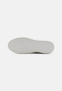 Suela de zapatilla blanca con un patrón de goma texturizado y acentos acanalados. Superficie lisa con un borde curvado, diseñada para ofrecer tracción y comodidad.