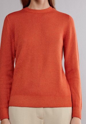Femme portant un pull orange uni à manches longues avec des poignets et un ourlet côtelés, associé à un pantalon beige sur un fond gris.