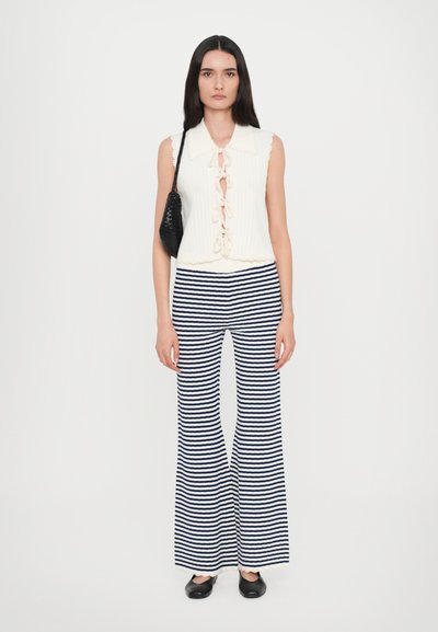 Top sans manches blanc à côtes avec un laçage sur le devant, associé à un pantalon évasé rayé bleu marine et blanc. Un sac noir et des chaussures plates complètent le look.