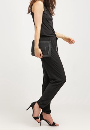 Jumpsuit noir sans manches avec une coupe décontractée, associé à des sandales à talons hauts noires et à une pochette structurée noire. Texture lisse.