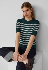 Grün gestreifter Kurzarmpullover mit weißen Horizontalstreifen. Gerippter Ausschnitt und Saum. Kombiniert mit dunklen Jeans und glänzenden burgunderfarbenen Schuhen.