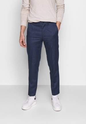 Mann trägt dunkelblaue maßgeschneiderte Hose, beige langärmlige Hemd und weiße Sneaker, steht vor einem schlichten hellen Hintergrund.