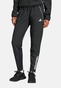 Pantalons de sport noirs avec une coupe fuselée, présentant des panneaux latéraux noirs contrastants et des rayures blanches Adidas le long des jambes.
