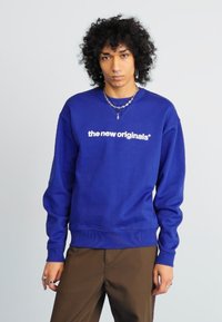 Persona con capelli ricci che indossa una felpa blu con il testo "the new originals", pantaloni marroni, gioielli argentati e un'espressione neutra.