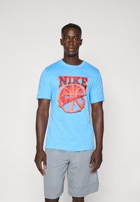 Lys blå Nike T-shirt med et stort orange basketballgrafik og logo. Korte ærmer og rund halsudskæring. Bæres med grå shorts.