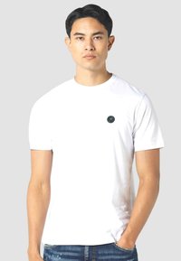 White cotton t-shirt with a crew neckline, short sleeves, and a small, round black logo on the chest. Paired with blue denim jeans.