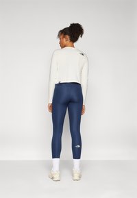 Hosszú ujjú, krémszínű felső, cropped szabással, amelyet magas derekú tengerészkék leggings-szel párosítanak, fehér logóval, amihez világos sportcipő illik.