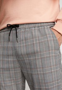 Person wearing gray plaid pants with black drawstring and a light pink sweater, hand resting in pocket.