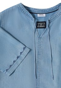 Tunique en denim bleu clair avec encolure en V, liens à cordon, manches 3/4 et broderie festonnée bleu foncé sur le poignet de la manche.