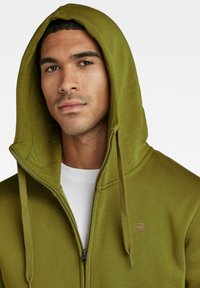 Felpa con zip di colore verde oliva, dotata di cappuccio con stringa, realizzata in un tessuto testurizzato, con tasche laterali e un piccolo logo sul petto.