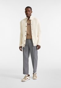 Cardigan voluminoso a coste color crema con colletto alto e bottoni marroni; indossato sopra una camicia beige e pantaloni grigi; completato da sneakers beige con dettagli.