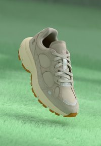 Beige sportskor tillverkade av läder och mocka, med rundad tå, ljusgrå accentfärger och en gummisula med orangea grepppunkter.