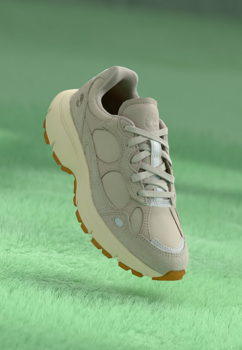Beige sportskor tillverkade av läder och mocka, med rundad tå, ljusgrå accentfärger och en gummisula med orangea grepppunkter.