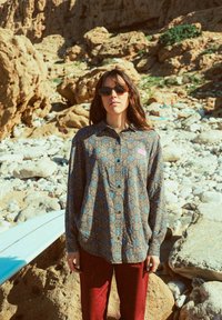 Femme portant une chemise à motifs et des lunettes de soleil se tient parmi un terrain rocheux avec une planche de surf reposant sur les rochers à proximité sous un soleil éclatant.