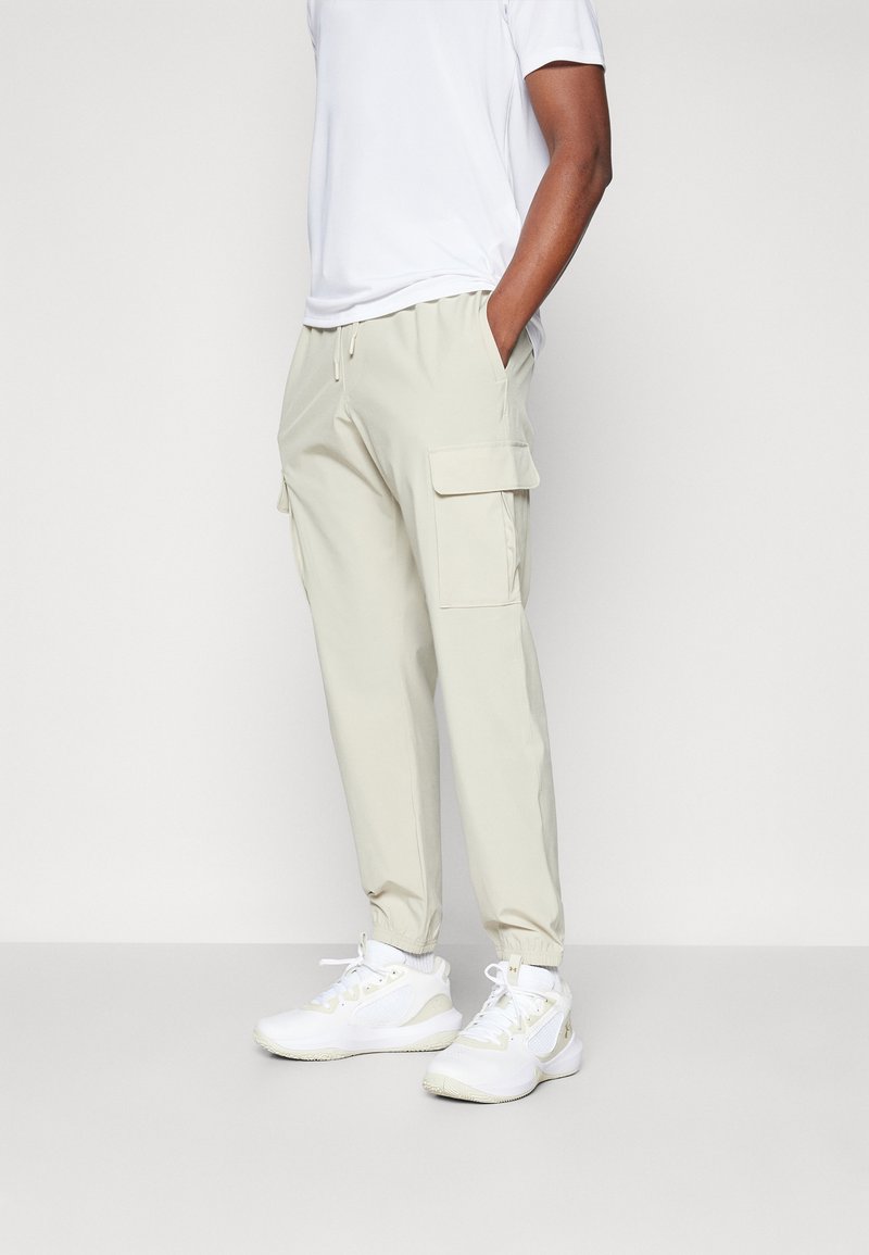 Pantalons cargo beiges avec poignets élastiques et taille à cordon de serrage, dotés de deux poches latérales et d'un tissu lisse et léger. Des baskets blanches complètent le look.
